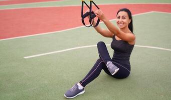 meisje atleet opleiding gebruik makend van trx Aan sport veld. gemengd ras jong volwassen vrouw Doen training met suspensie systeem. gezond levensstijl. uitrekken buitenshuis speelplaats. foto