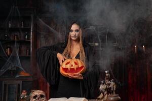 halloween concept. zwart heks Holding halloween pompoen met gesneden lachend gezicht in hand- staand donker kamer foto