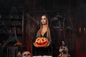 halloween concept. zwart heks Holding halloween pompoen met gesneden lachend gezicht in hand- staand donker kamer foto