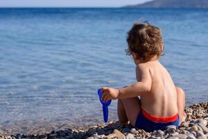 terug visie van een weinig bruinen jongen zittend Aan de kust Aan een heet zomer vakantie dag. hij is Holding een klein blauw Schep. visie van kristal schoon water oppervlakte mediterraan, agia fotini strand chios Griekenland foto