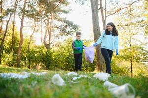 vrouw vrijwilliger en weinig jongen plukken omhoog de plastic vuilnis en zetten het in biologisch afbreekbaar vuilniszak buitenshuis. ecologie, recycling en bescherming van natuur concept. milieu bescherming. foto