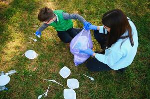 vrouw vrijwilliger en weinig jongen plukken omhoog de plastic vuilnis en zetten het in biologisch afbreekbaar vuilniszak buitenshuis. ecologie, recycling en bescherming van natuur concept. milieu bescherming. foto