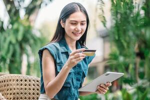 jong vrouw Holding credit kaart en gebruik makend van Tablet. een glimlachen jong vrouw bezighoudt met een tablet temidden van levendig groen gebladerte. online winkelen, e-commerce, internet bankieren, uitgeven geld. foto