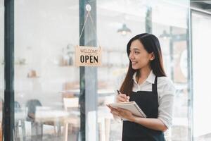 een glimlachen zakenvrouw door de ​ Welkom open'' teken van haar winkel duurt notities, voorbereidingen treffen voor de dag taken in haar helder en uitnodigend cafe, belichamen de geest van ondernemend gereedheid. foto