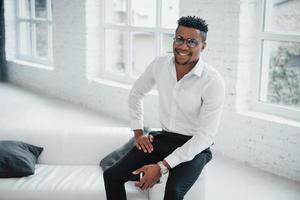 stijlvolle afro-amerikaanse man in kantoorkleding en een bril die aan de rand van de bank zit en glimlacht