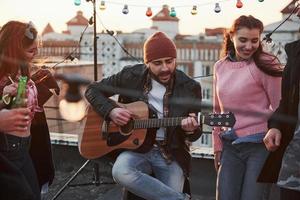 gitarist zit en zingt voor zijn vrienden op het dak met decoratieve gekleurde gloeilampen foto