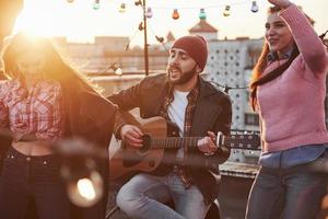 voel het ritme. gitarist zit en zingt voor zijn vrienden op het dak met decoratieve gekleurde gloeilampen foto