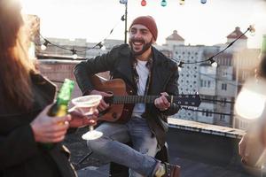 foto van jonge, bebaarde aantrekkelijke gitarist die op het dak voor zijn vrienden zingt