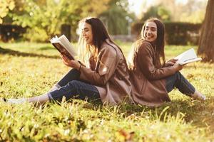 lachende brunette tweelingmeisjes die rug aan rug op het gras zitten en plezier hebben met benen licht gebogen in de knieën, met bruine boeken in handen, casual jas dragend in herfstpark op onscherpe achtergrond foto