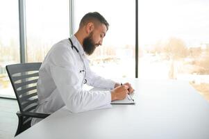 jong 30 jaar oud knap aangenaam Arabisch mannetje dokter in wit jas, poseren Bij camera binnenshuis. foto