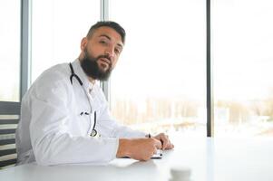 jong 30 jaar oud knap aangenaam Arabisch mannetje dokter in wit jas, poseren Bij camera binnenshuis. foto