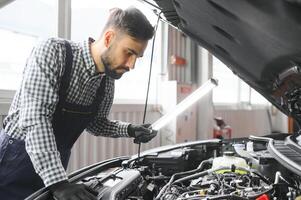 professioneel monteur is werken Aan een auto in een auto onderhoud. klusjesman in veiligheid bril is vaststelling de motor Aan een voertuig. specialist draait los bouten met een ratel. modern schoon werkplaats foto