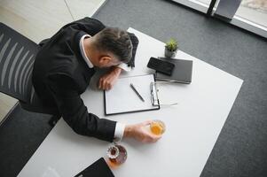 dichtbij omhoog van benadrukt zakenman Holding een glas van whisky hij slapen en gegevens grafieken, zaken document Bij kantoor bureau. alcohol verslaving en dronken zakenman concept foto