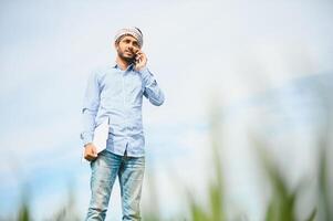 portret van gelukkig jong Indisch Mens boer staand in landbouw land- omringen door Bijsnijden Holding smartphone pratend naar iemand, glimlachen dorp mannetje met mobiel telefoon in veld, technologie concept foto