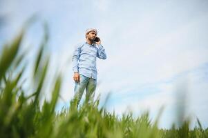 portret van gelukkig jong Indisch Mens boer staand in landbouw land- omringen door Bijsnijden Holding smartphone pratend naar iemand, glimlachen dorp mannetje met mobiel telefoon in veld, technologie concept foto