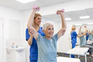 Oder oud vrouw aan het doen fysiotherapie met ondersteuning van fysiotherapeuten . senior ouderen vrouw zittend in kliniek gebruik makend van halters training oefening voor geduldig met verzorger in verpleging zorg. foto