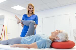 dichtbij omhoog van vrouw fysiotherapeut is helpen senior ouderling vrouw uitrekken haar hamstring en aan het doen dij of been revalidatie in oefening kamer - ze is aan het liegen Aan massage bed foto