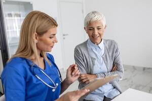 jong vrouw dokter of gp in wit medisch uniform raadplegen senior vrouw geduldig in privaat ziekenhuis. vrouw therapeut spreken praten met vrouw cliënt Aan overleg in kliniek. foto