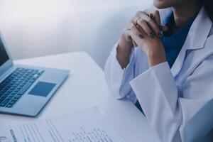 dokter en geduldig zittend Bij de bureau in kliniek kantoor. de focus is Aan vrouw arts handen vulling omhoog de geneesmiddel geschiedenis Vermelding het formulier, dichtbij omhoog. geneeskunde concept foto