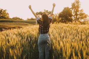 jong mooi vrouw in rood zomer jurk en rietje hoed wandelen Aan geel boerderij veld- met rijp gouden tarwe genieten van warm avond. foto