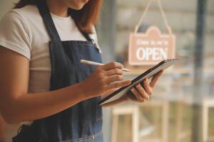 startup succesvolle kleine ondernemer mkb schoonheid meisje staan met tablet smartphone in coffeeshop restaurant. portret van aziatische tan vrouw barista café-eigenaar. MKB ondernemer verkoper bedrijfsconcept foto