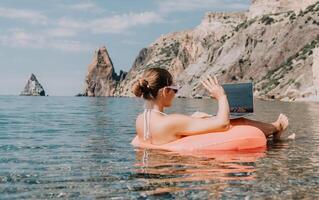 vrouw freelancer werken Aan laptop zwemmen in zee Aan roze opblaasbaar ring. gelukkig toerist in zonnebril drijvend Aan opblaasbaar donut en werken Aan laptop computer in kalmte oceaan. afgelegen werken overal foto