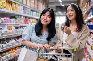 twee jong Aziatisch Dames winkel samen in een kruidenier op te slaan, gefocust Aan selecteren items van de planken. foto