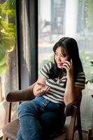 een positief Aziatisch vrouw is pratend Aan de telefoon met iemand terwijl zittend in een koffie winkel. foto