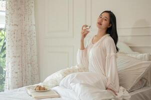 een aantrekkelijk Aziatisch vrouw in lang jurk pyjama is toepassen parfum na ontwaken omhoog Aan haar bed. foto