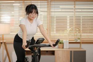 een gezond, fit Aziatisch vrouw in sportkleding is aan het doen cardio Bij huis, oefenen Aan een geschiktheid fiets. foto