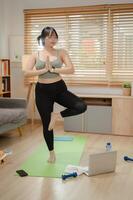 een sterk Aziatisch vrouw in sportkleding is beoefenen yoga Bij huis, aan het doen de boom houding Aan een yoga mat. foto