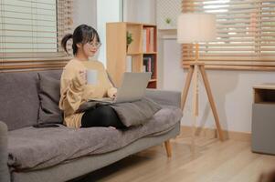 een aantrekkelijk Aziatisch vrouw is werken van een afstand Bij huis, nippen koffie, en werken Aan haar laptop. foto