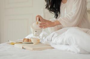 een vrouw in lang jurk pyjama gieten thee van een mooi keramisch theepot, hebben ontbijt in bed foto