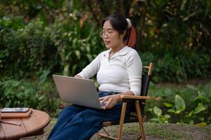 een jong Aziatisch vrouw is werken van een afstand in haar achtertuin of Bij een buitenshuis cafe, gebruik makend van haar laptop. foto