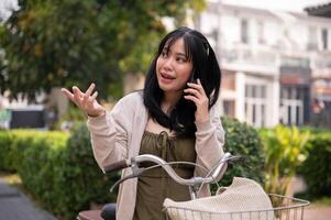 een mooi, gelukkig jong Aziatisch vrouw is pratend Aan de telefoon terwijl rijden een fiets in de stad. foto