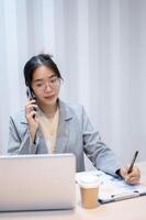 een zakenvrouw is pratend Aan de telefoon, nemen notities, en lezing informatie Aan haar laptop. foto
