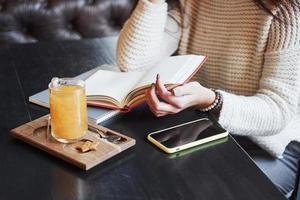 bijgesneden close-up foto van vrouw lees het boek in restaurant met gele drank en smartphone op tafel
