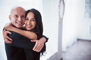 omhelzen elkaar en glimlachen. portret van gelukkige paar binnenshuis. kale man en brunette vrouw staat in de witte kamer foto