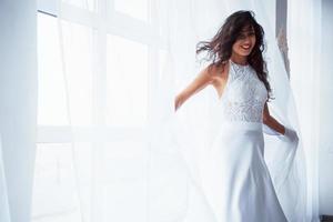 glanzende foto. mooie vrouw in witte jurk staat in witte kamer met daglicht door de ramen
