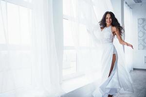 mooie vrouw in witte jurk staat in witte kamer met daglicht door de ramen