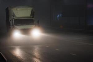 een vrachtauto met koplampen het rijden Aan een mistig snelweg Bij middernacht foto