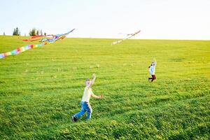 glimlachen meisje en broer jongen rennen met vliegend kleurrijk vliegers Aan de hoog gras weide. gelukkig kinderjaren momenten foto