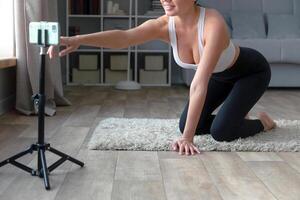 vrouw zittend Aan de verdieping in yoga broek opname een online geschiktheid met haar telefoon en statief, glimlachen Bij de camera in een leven kamer instelling foto