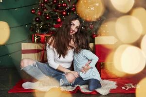 meisje geeft een kus. vrolijke moeder en dochter zitten in de buurt van de kerstboom die erachter zit. schattig portret foto