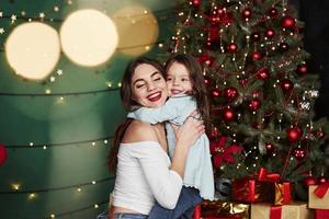 geweldige vakantiestemming. je kunt het zien aan hun glimlach. vrolijke moeder en dochter knuffelen elkaar in de buurt van de kerstboom die erachter zit. schattig portret foto