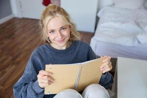 portret van lachend, charismatisch jong vrouw, schrijven naar beneden notities, maken plannen en zetten het in planner, Holding logboek, zittend in slaapkamer en op zoek gelukkig Bij camera foto