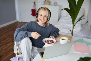 beeld van gelukkig vrouw zittend in een kamer, aan het kijken interessant TV tonen of film Aan laptop, gebruik makend van schreeuwen onderhoud, vervelend koptelefoon, aan het eten toetje en drinken thee foto