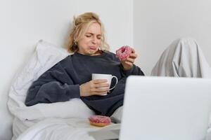 portret van vrouw aan het kijken verdrietig film Aan laptop, huilen en afvegen tranen uit, aan het eten donut en drinken thee foto