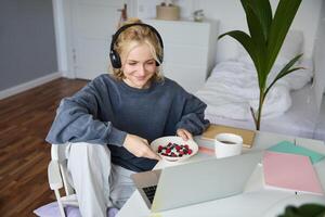 portret van glimlachen jong vrouw, aan het kijken TV tonen in koptelefoon, aan het eten ontbijt en op zoek Bij laptop scherm foto