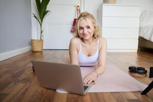 portret van jong geschiktheid vrouw, aan het doen training Bij huis, op zoek Bij haar Sportschool instructeur Aan laptop scherm, aan het doen online opleiding sessie foto